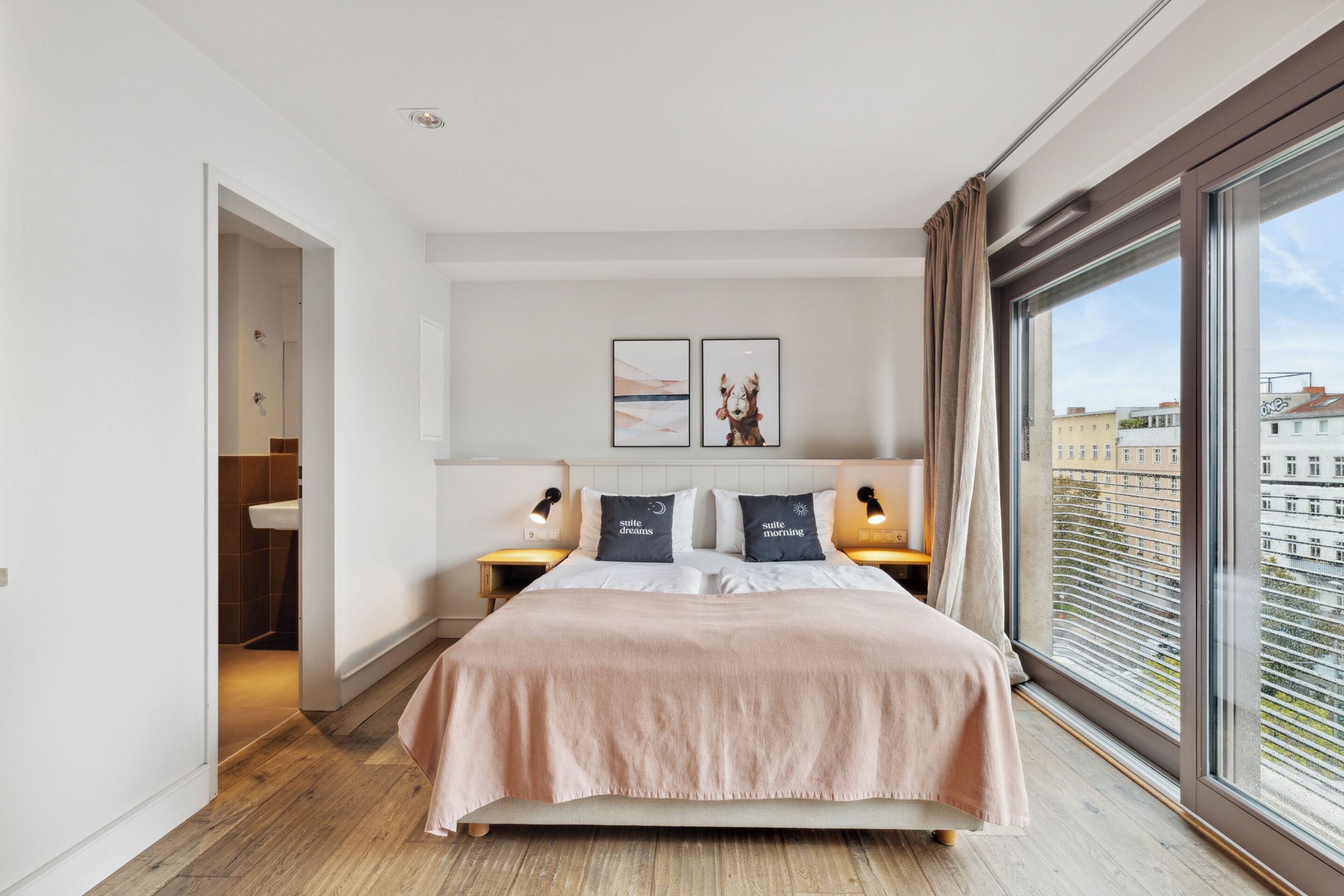 Une chambre moderne avec un grand lit, deux tables de chevet avec des lampes, une vue sur les immeubles à travers une grande fenêtre et des œuvres d’art accrochées au mur.