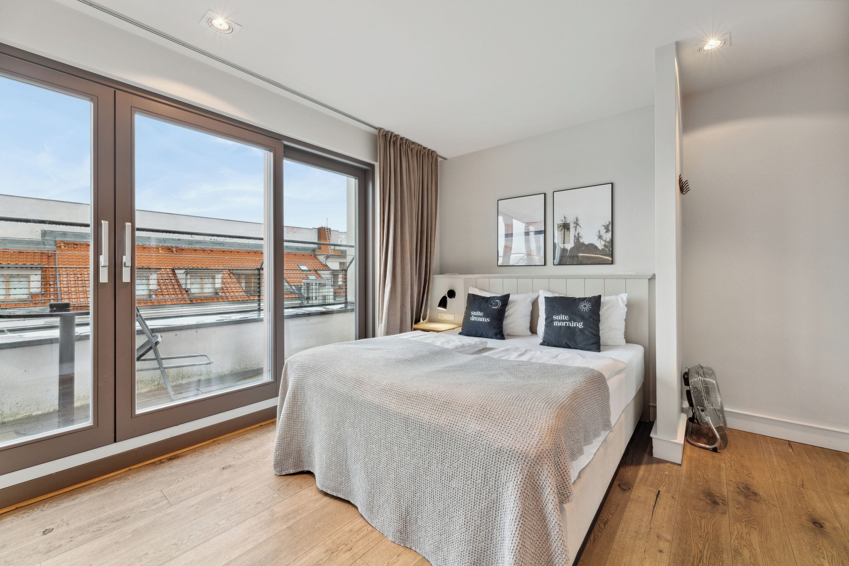 A modern bedroom with a large window, a bed with a grey bedspread, two bedside tables, and a radiator.