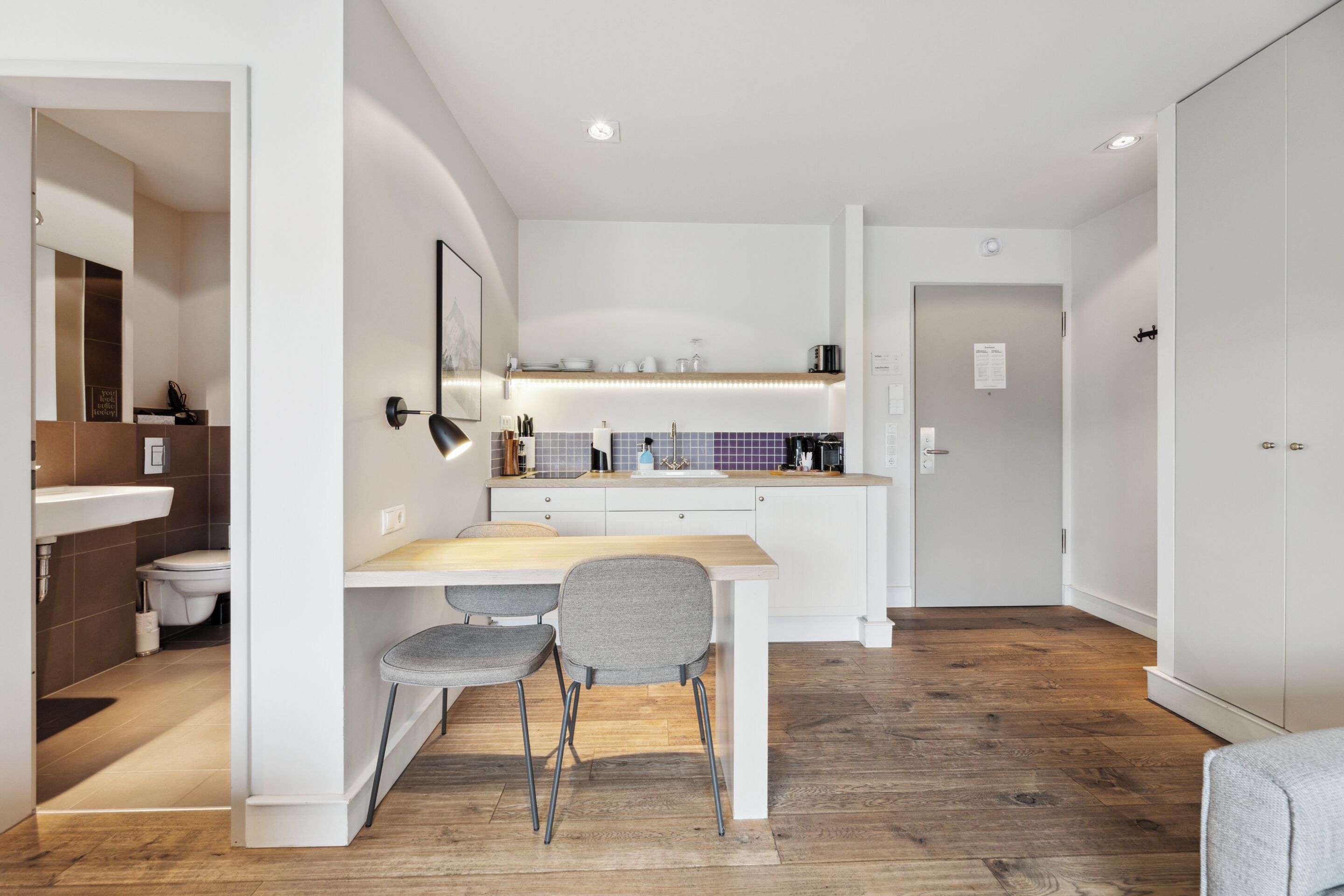 A modern kitchen with white cabinetry, a wooden countertop, and a built-in stove. There is a dining area with a table and two chairs. The room has a wooden floor and a large mirror on the wall.