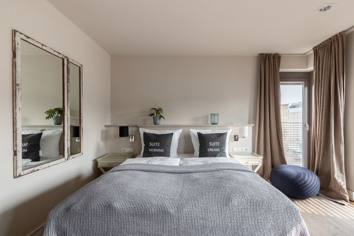 Une chambre d’hôtel avec un grand lit, deux tables de chevet, un miroir, une fenêtre avec des rideaux et une vue sur les bâtiments.