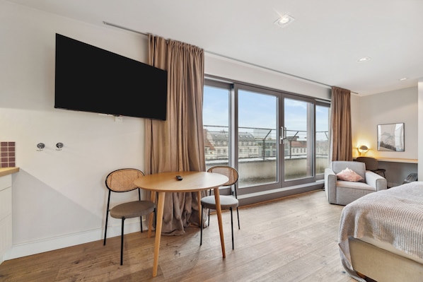 Una habitación de hotel moderna con un ventanal grande, un televisor de pantalla plana, una zona de comedor con mesa y sillas, una cama y un sofá.