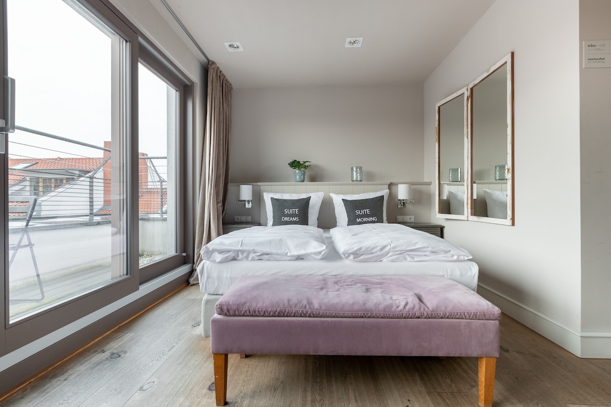 Une chambre moderne avec une grande fenêtre, un lit recouvert d’une literie blanche, une table de chevet avec une lampe et un miroir au mur.