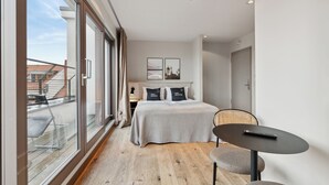 Een moderne hotelkamer met een groot bed, een klein tafeltje en uitzicht op de stad door grote ramen.