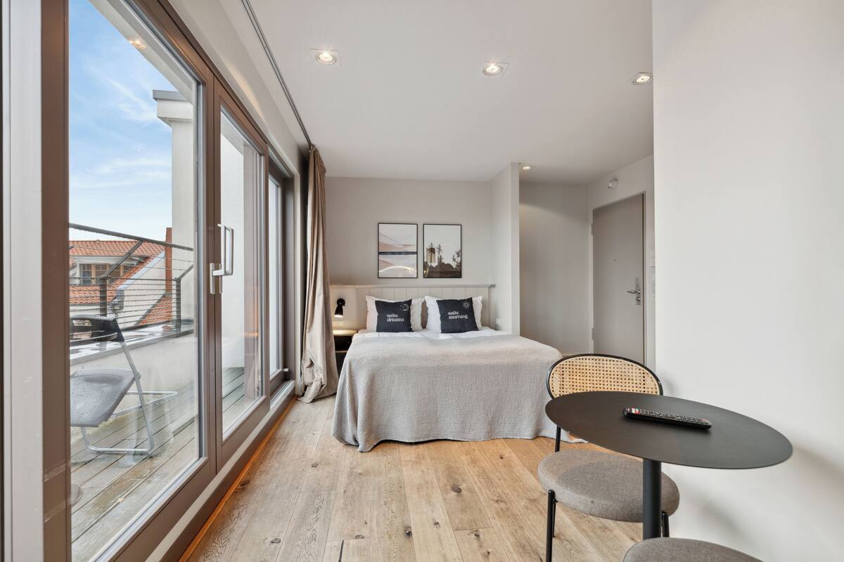 Une chambre d’hôtel moderne, dotée d’un grand lit, d’une petite table et offrant une vue sur la ville grâce à de grandes fenêtres.