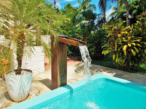 A swimming pool with a waterfall feature, surrounded by lush greenery and a white brick wall.