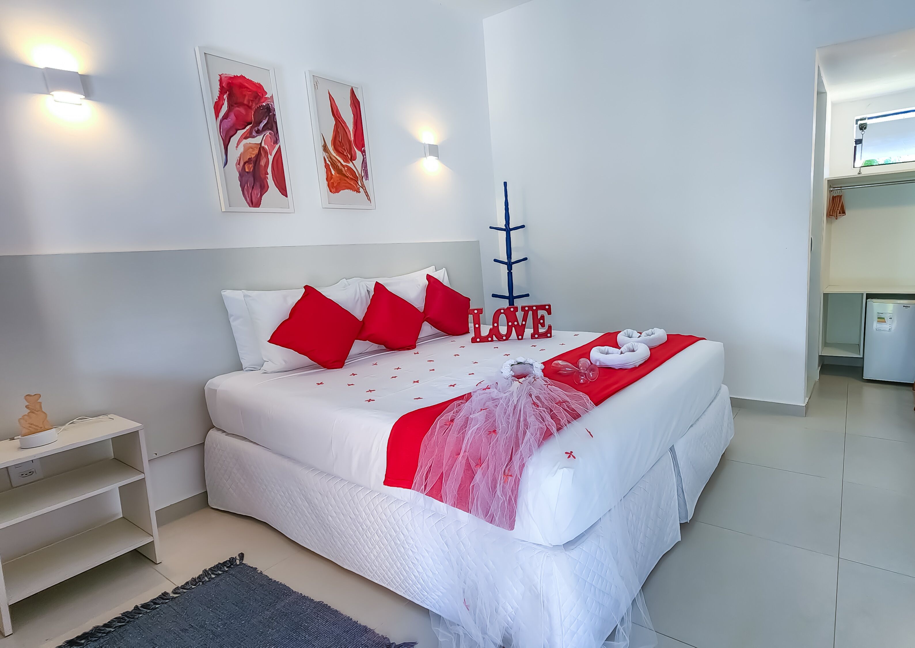 A modern bedroom with a large bed, red pillows, a 'LOVE' sign, and a small refrigerator in the corner.