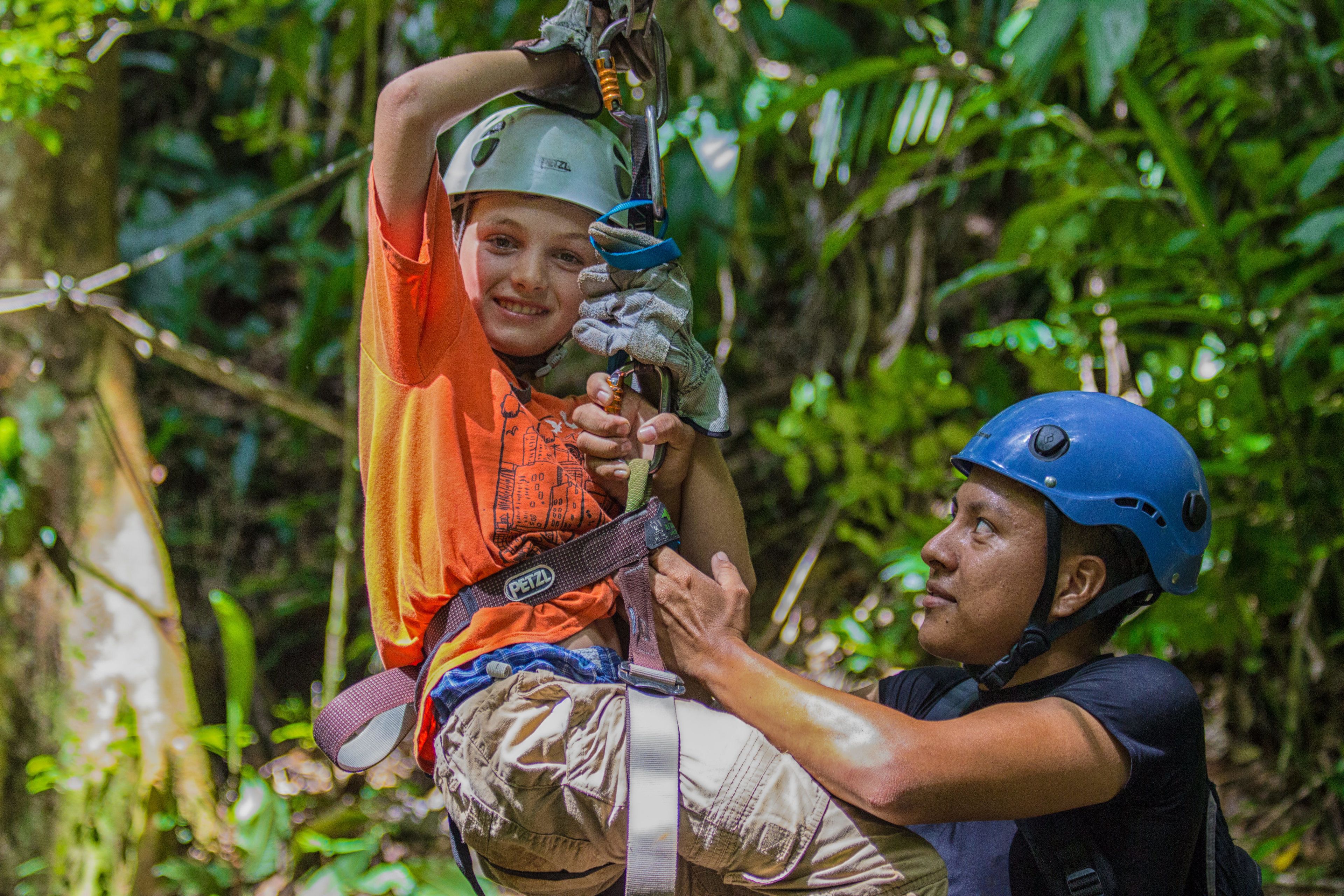 A person in a helmet and harness being assisted by another person in a forested area.