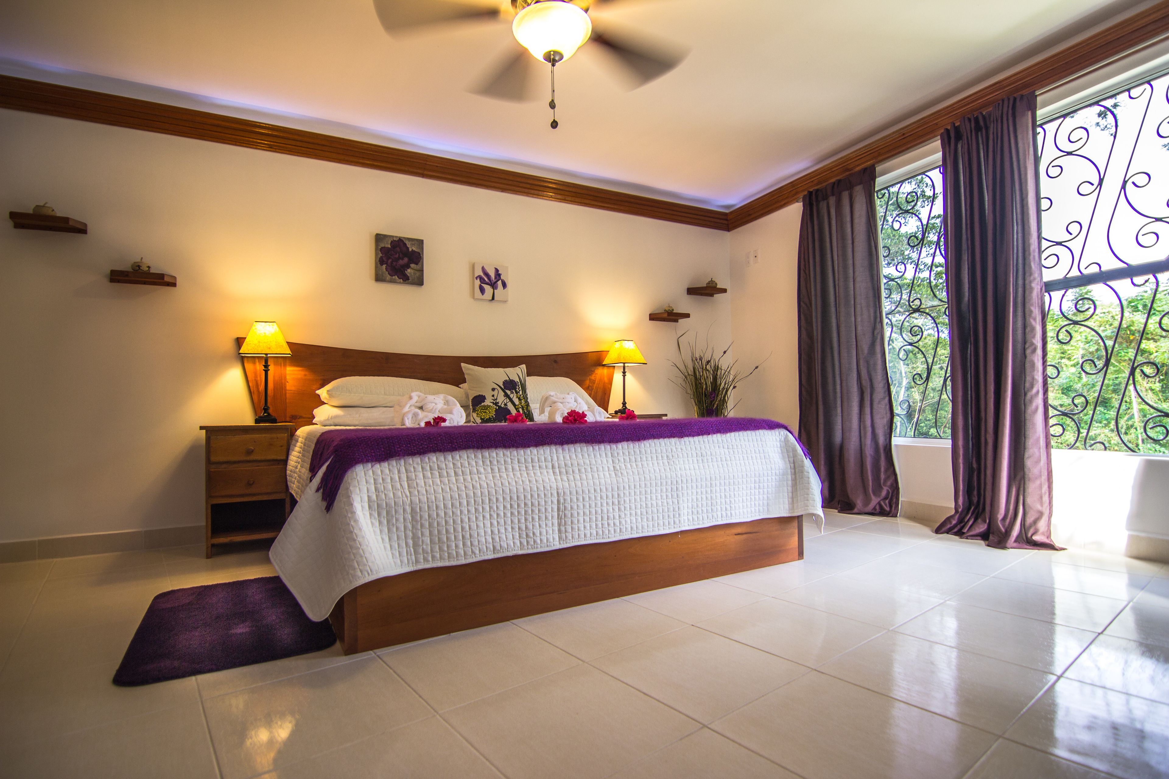 A bedroom with a large bed, two bedside tables with lamps, a ceiling fan, and a view of greenery through a window with decorative iron bars.
