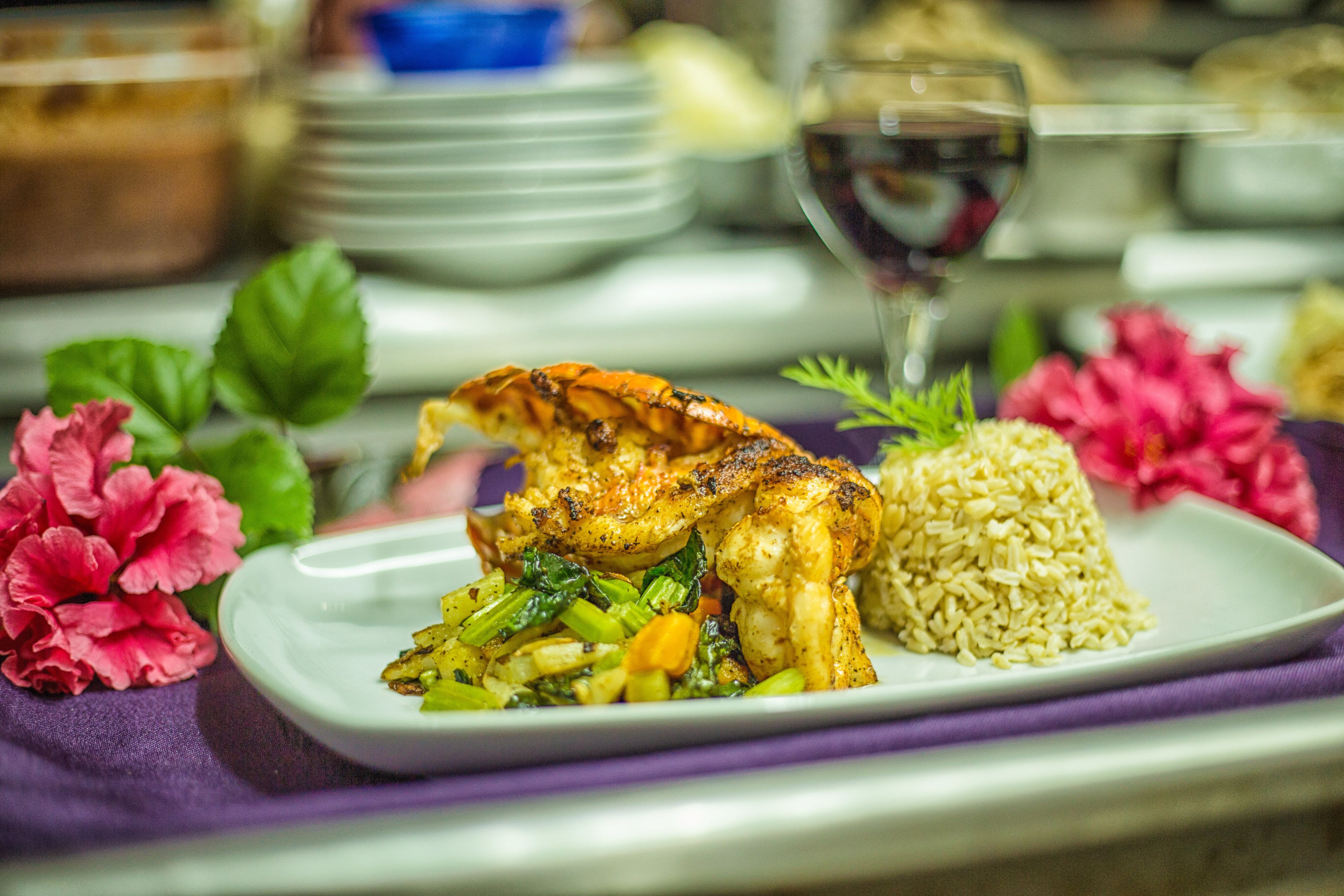 Prato de comida com arroz e legumes, um copo de vinho tinto e flores cor-de-rosa sobre uma mesa.