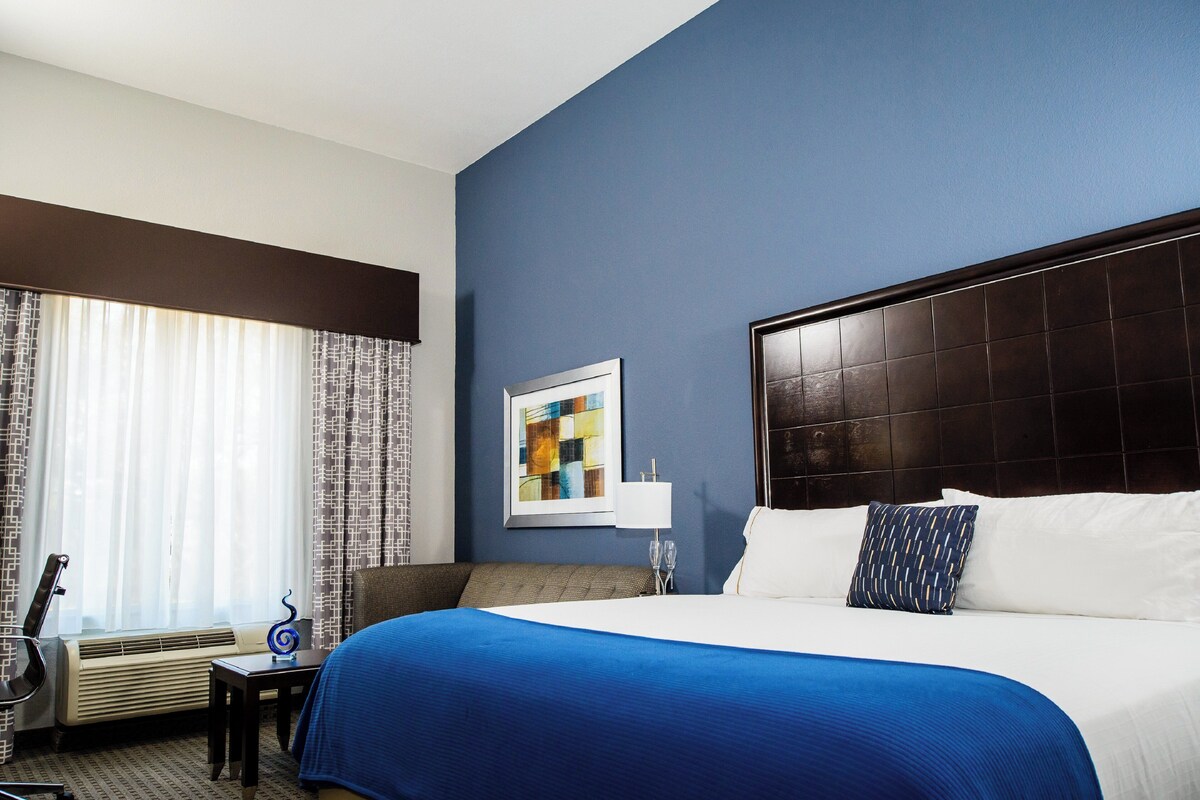 A hotel room with a large bed, a desk with a chair, a window with curtains, and a wall-mounted artwork.