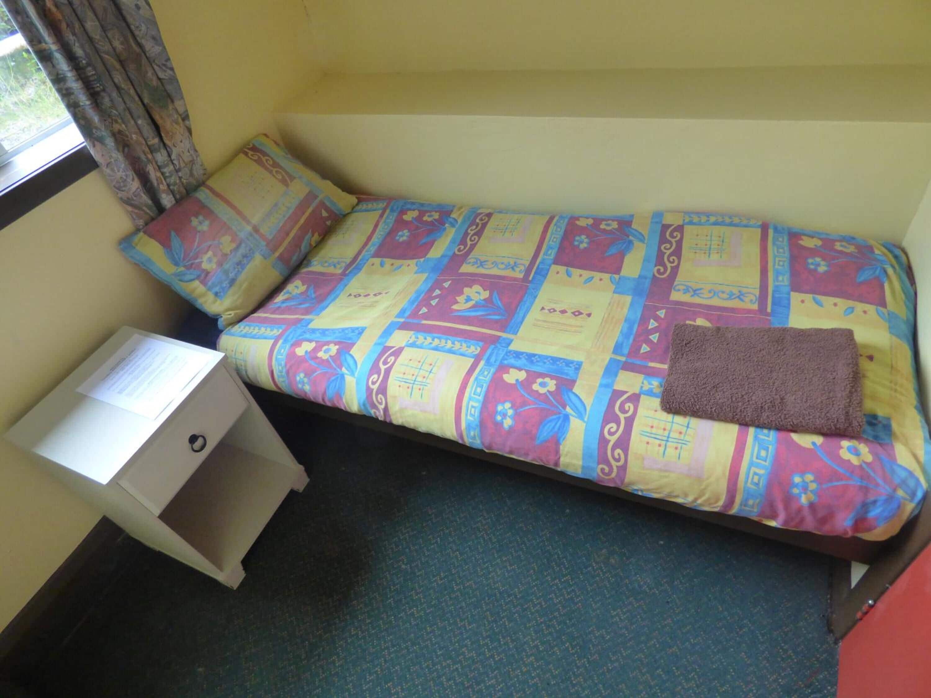 A small, single-bed room with a colorful quilted bedspread, a brown towel, and a white nightstand with a lamp.