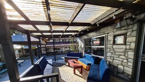 A covered outdoor seating area with wooden benches and blue cushioned sofas, stone walls, and a view of a building in the background.
