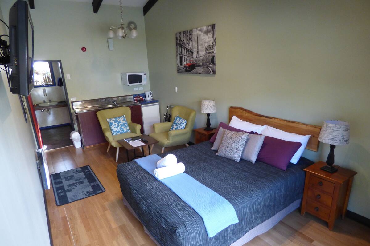 A hotel room with a bed, two chairs, a desk, a microwave, a sink, and a painting on the wall.
