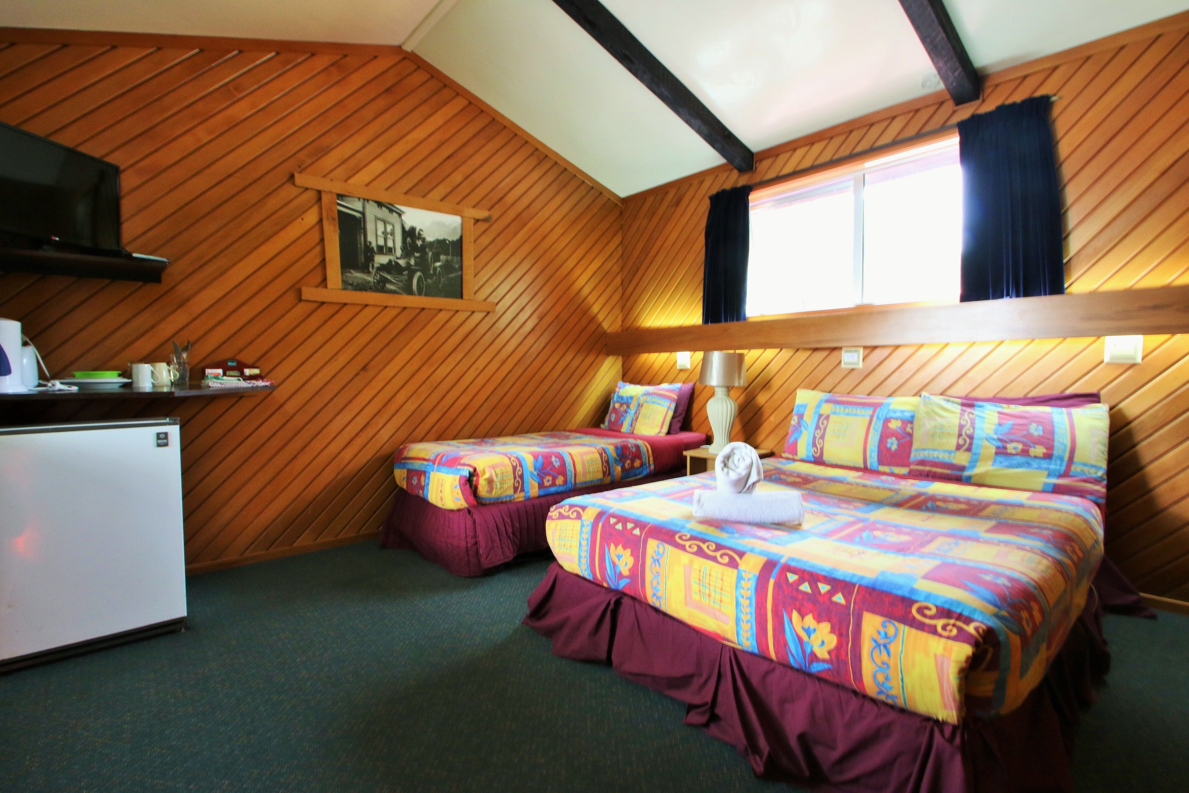 A room with wooden panel walls, two beds with colorful bedding, a small table with a lamp, and a refrigerator.