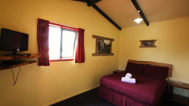 Una habitación de hotel con cama, televisor sobre un soporte, ventana con cortinas rojas y un cuadro colgado en la pared.