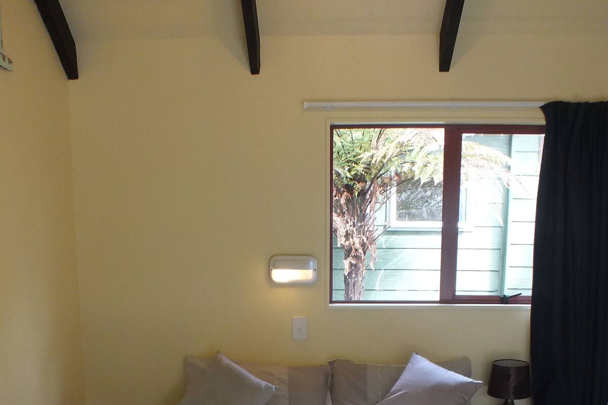 A neatly made bed with a rolled towel, a window with a view of greenery, and a bedside lamp.