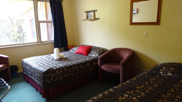 A room with two beds, a chair, a window with curtains, a wall-mounted mirror, and a small shelf with a picture. - Glow Worm Accommodation - Hostel (Franz Josef Glacier)