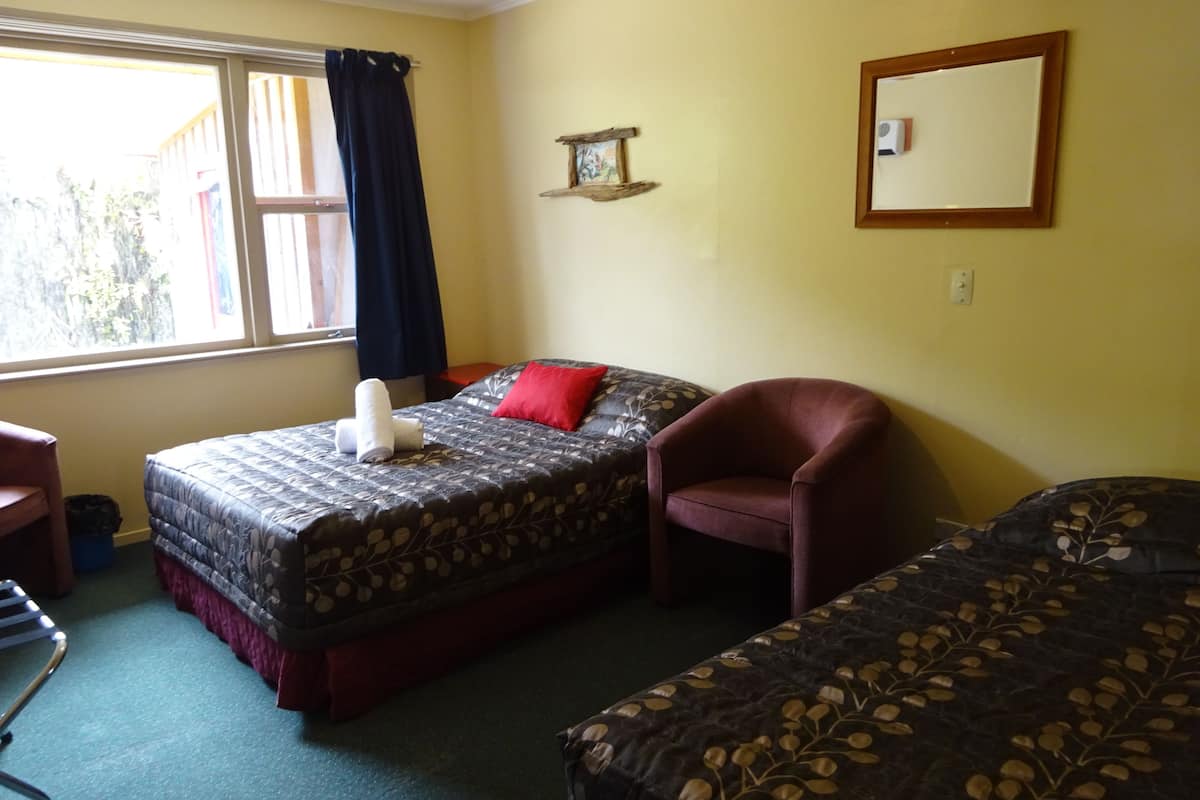 A room with two beds, a chair, a window with curtains, a wall-mounted mirror, and a small shelf with a picture.