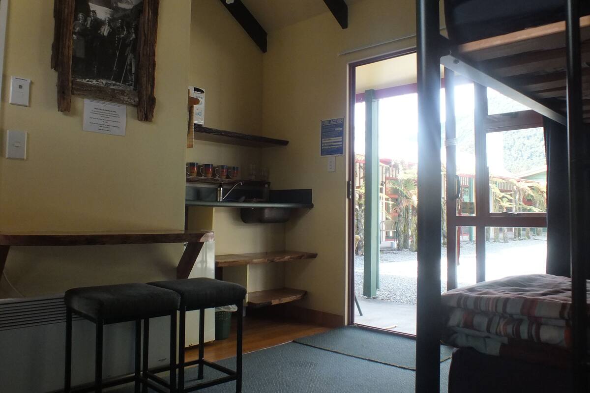 A room with a bed, a bench, a sink, and a door leading outside.