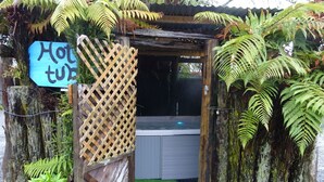 A rustic hot tub area with a wooden lattice entrance, surrounded by lush ferns and a sign indicating the temperature. - Glow Worm Accommodation - Hostel (Franz Josef Glacier)