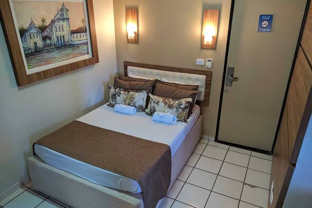 A bedroom with a bed, pillows, a blanket, a wall-mounted painting, and a door with a sign.