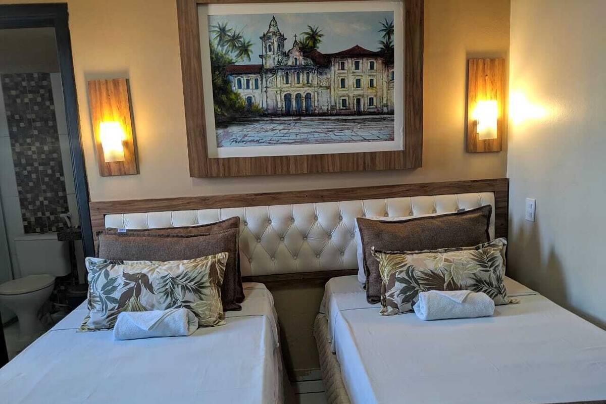 A hotel room with two beds, a large painting of a building, and wall-mounted lights.