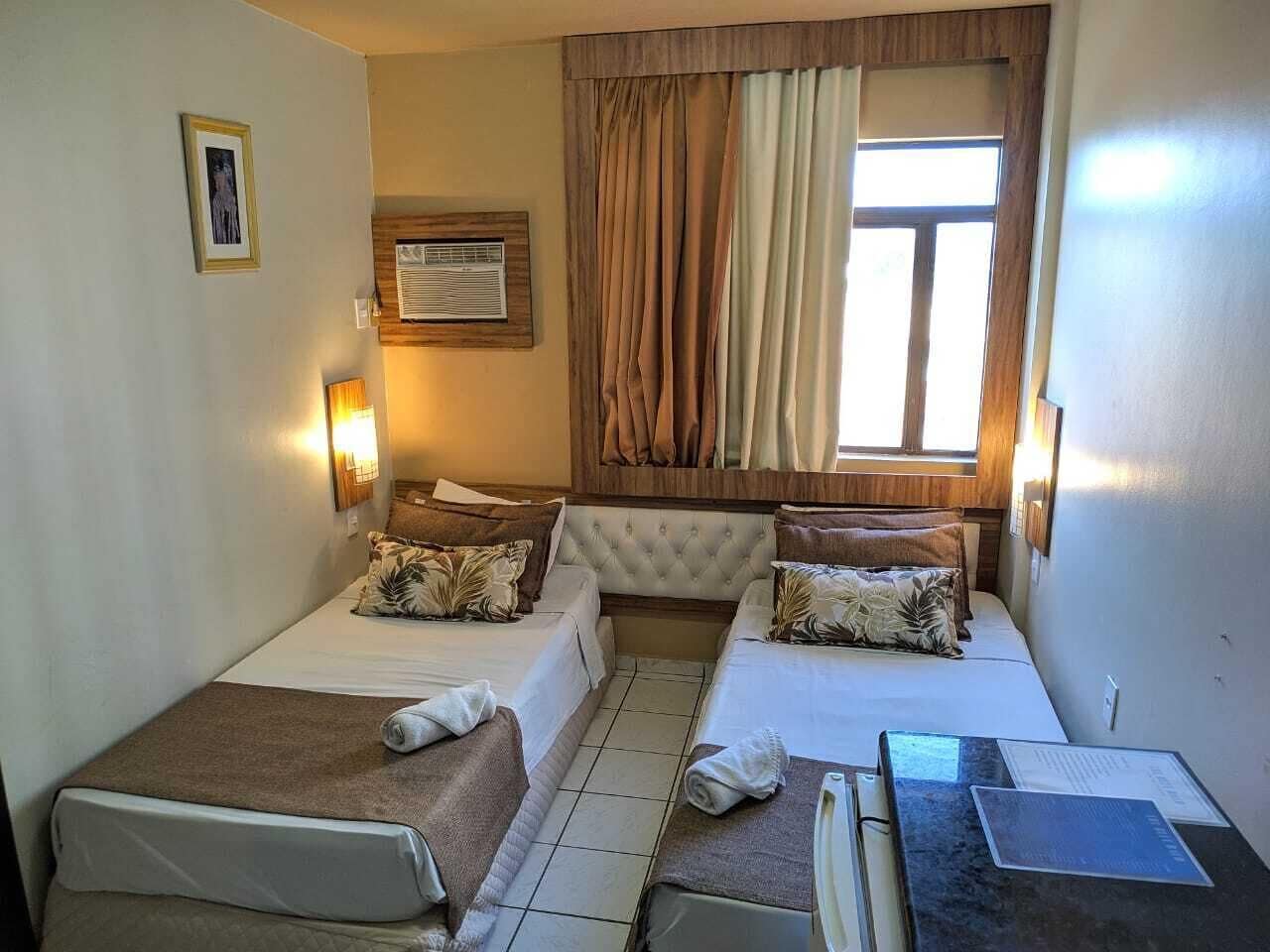 Une chambre d’hôtel de taille modeste, comprenant deux lits, une petite table, une fenêtre avec des rideaux, un climatiseur fixé au mur et un tableau encadré.