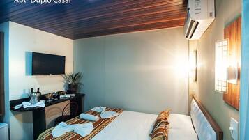 Quarto de hotel com cama, escrivaninha, televisão de tela plana, planta em vaso e aparelho de ar condicionado.