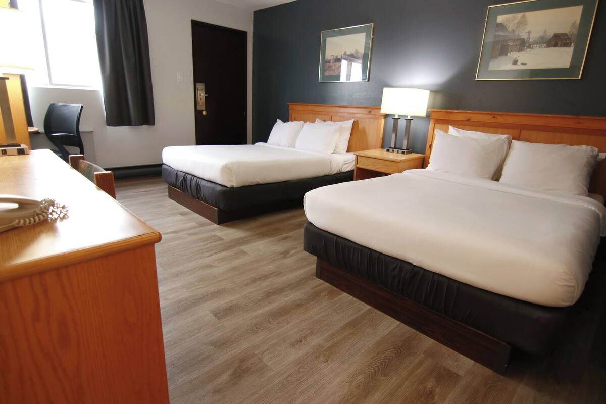 A hotel room with two beds, a wooden desk, a chair, a nightstand with a lamp, and a framed picture on the wall.