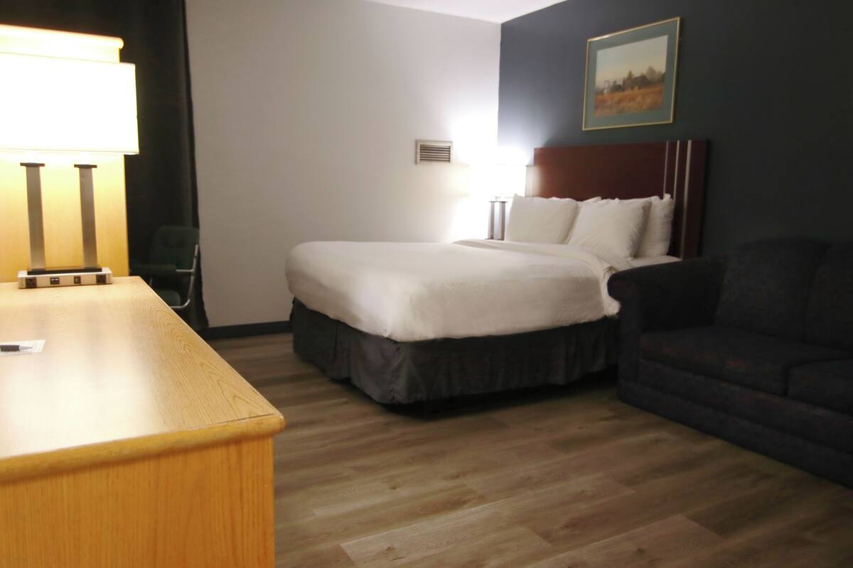 A hotel room with a bed, a desk, a chair, a couch, a lamp, and a painting on the wall.