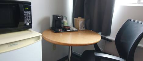 Habitación de hotel pequeña y funcional, con microondas, una mesita con cafetera, una silla y ventana con cortinas.