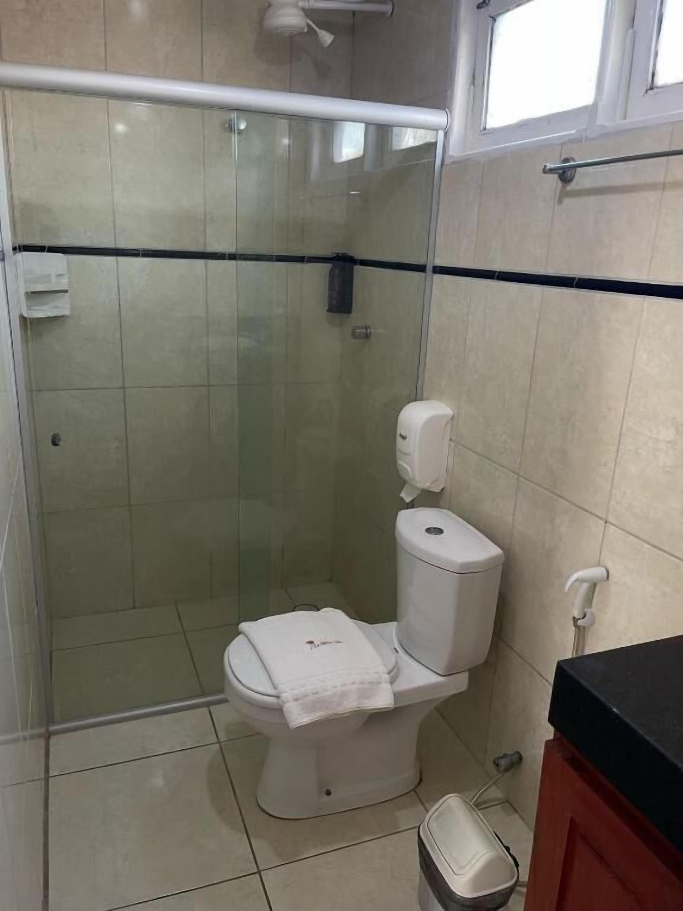 A bathroom with a glass-enclosed shower, a toilet with a paper towel holder, and a trash bin.