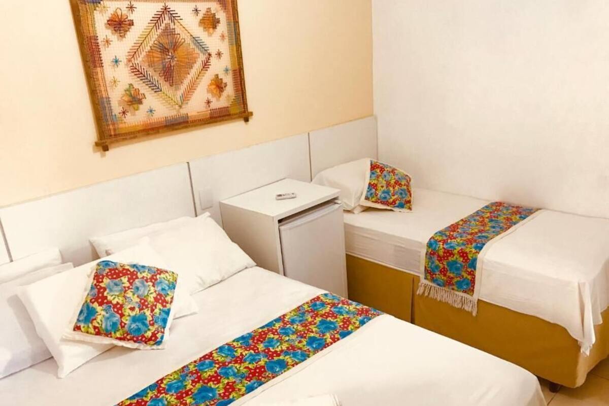 Habitación de hotel con dos camas, cada una con ropa de cama blanca y mantas decorativas con motivos florales. Se observa una mesita de noche con una lámpara y una obra de arte colgada en la pared sobre las camas.