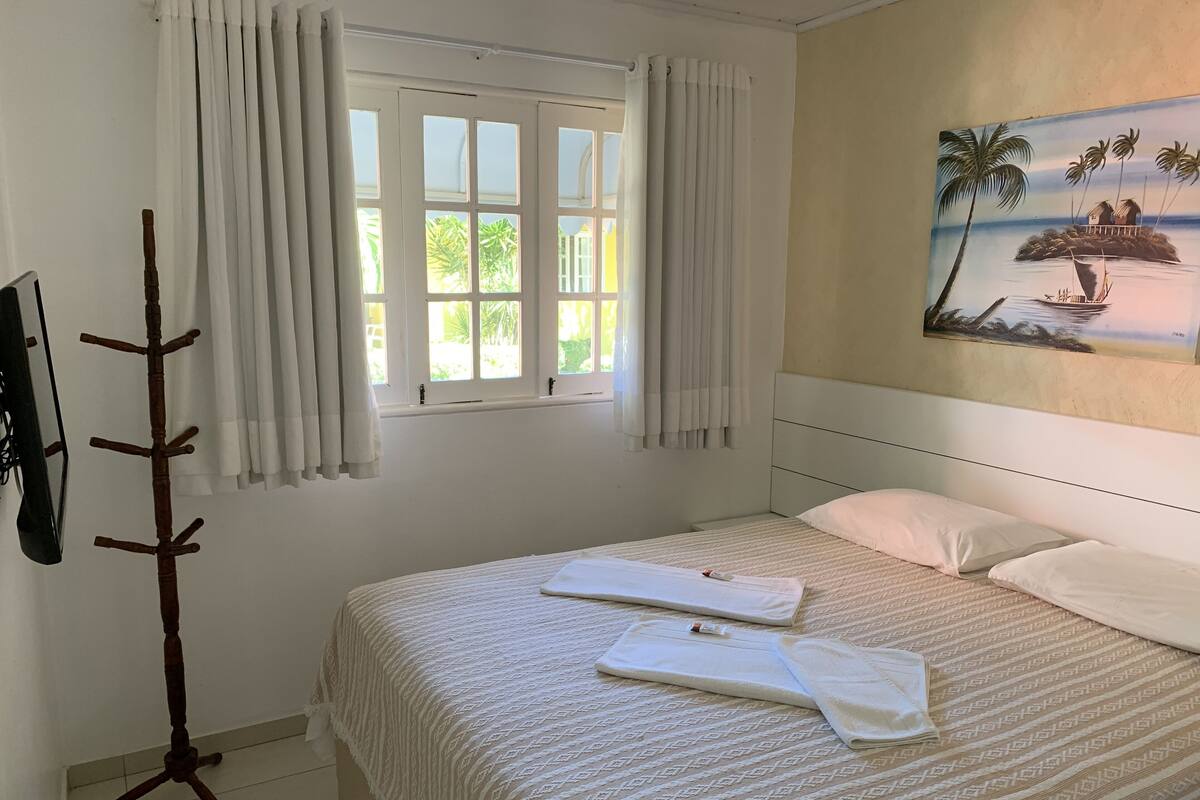 Una cama bien hecha con una colcha estampada y dos almohadas, una ventana con cortinas, un perchero y un cuadro con un paisaje tropical en la pared.