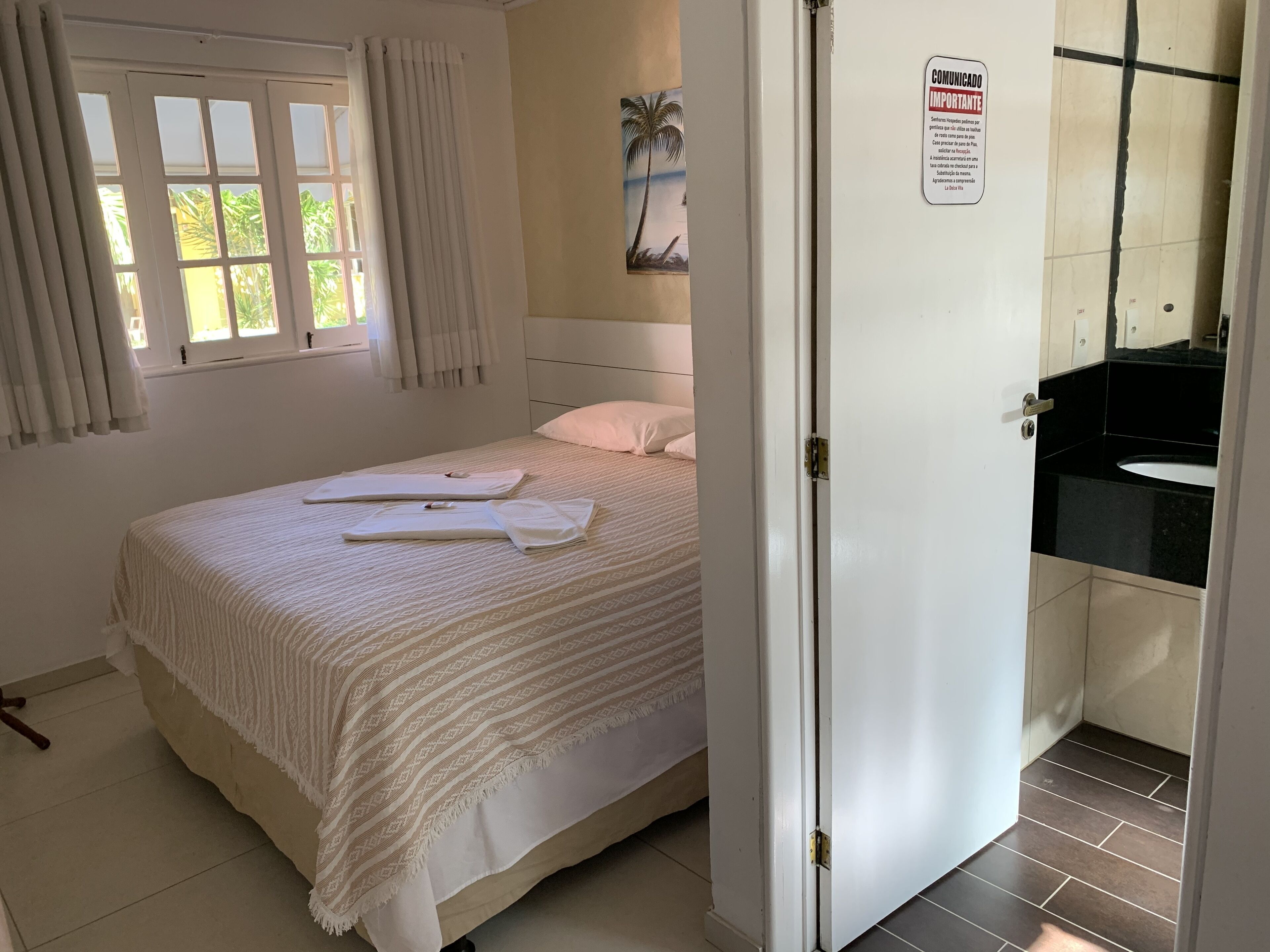Un lit bien fait, recouvert d’une courtepointe blanche et agrémenté d’oreillers, une fenêtre avec des rideaux, un tableau accroché au mur et une salle de bain visible à travers une porte ouverte.