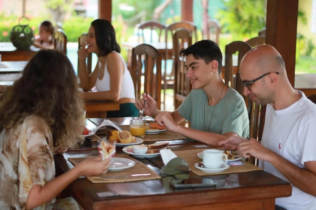 Un groupe de personnes est assis à un café en plein air, autour de tables et de chaises en bois, dans un cadre verdoyant.
