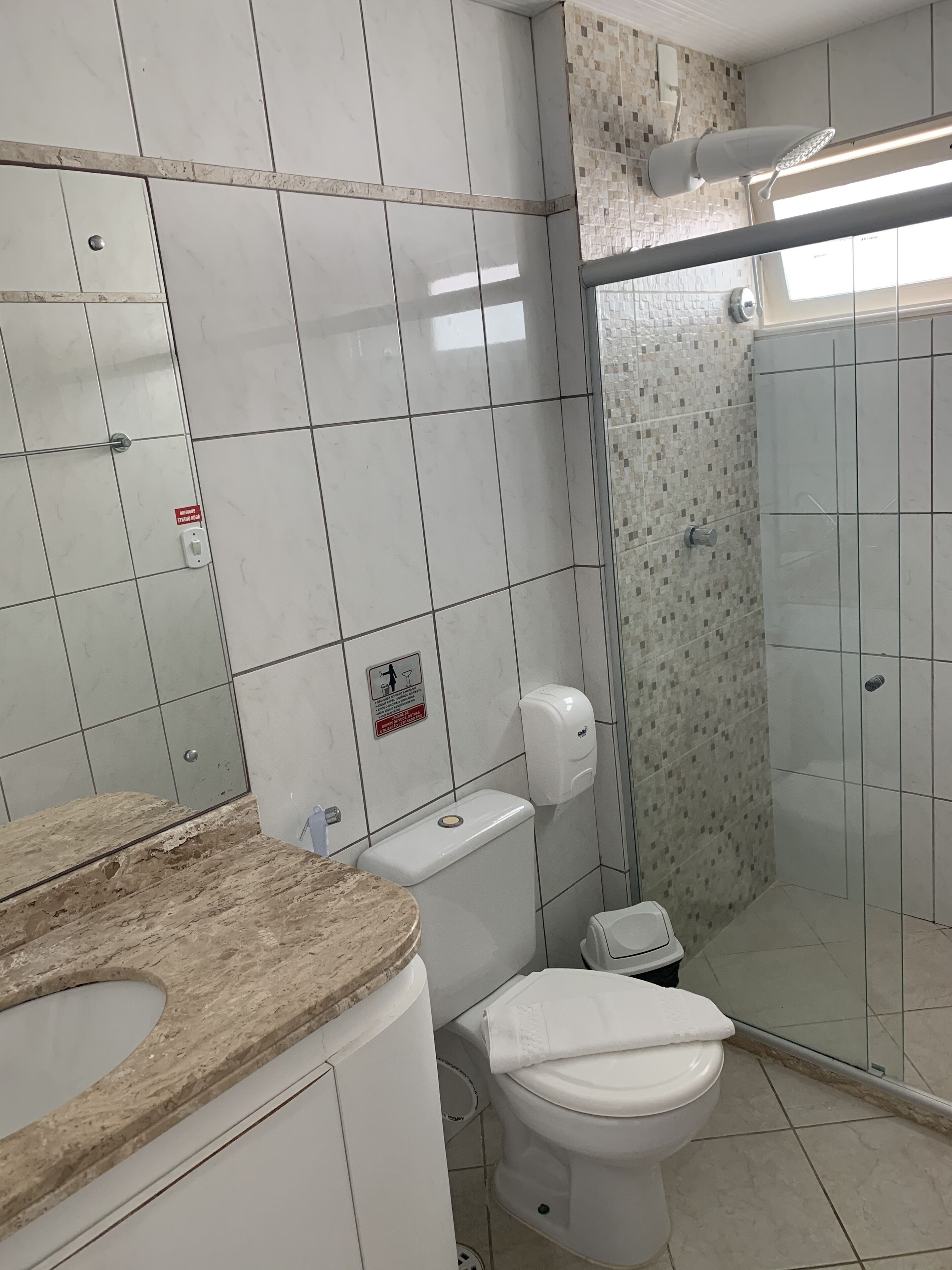 Een badkamer met witte betegelde wanden, een marmeren aanrecht, een toilet, een glazen douchecabine en een handdroger.
