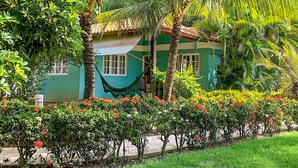 A green house with a hammock, surrounded by lush greenery and flowering plants.
