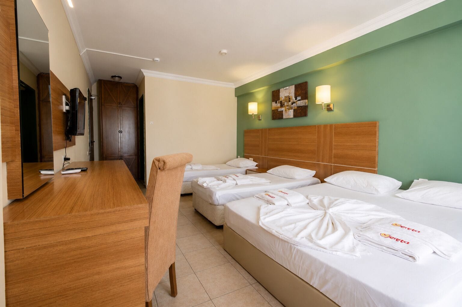 A hotel room with a wooden desk, a television, a bed with neatly made white linens, and a green accent wall.