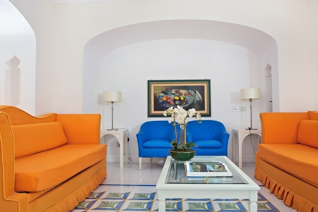 A room with two orange sofas, a blue armchair, a white coffee table with a vase of flowers, and a framed painting on the wall.