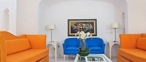A room with two orange sofas, a blue armchair, a white coffee table with a vase of flowers, and a framed painting on the wall.