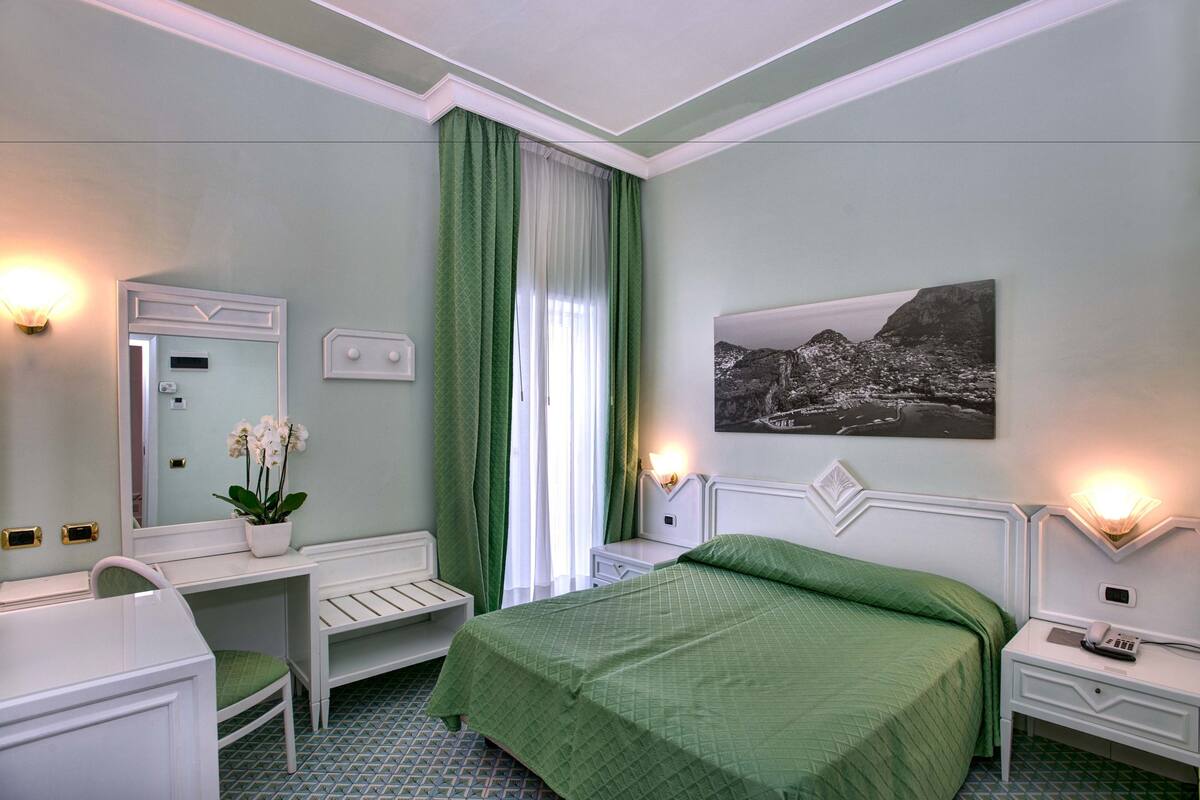 Ein Hotelzimmer mit Bett, Schreibtisch, Stuhl, Nachttisch, Lampe, Spiegel, Fenster mit Vorhängen und einem Wandbild.