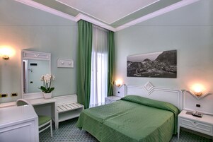 Habitación de hotel con cama, escritorio, silla, mesita de noche, lámpara, espejo, ventana con cortinas e imagen colgada en la pared.