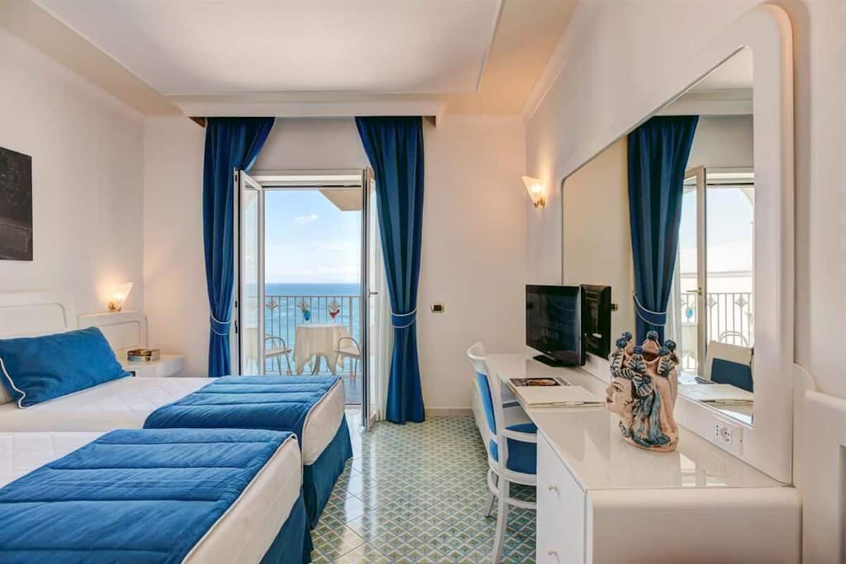 Ein Hotelzimmer mit zwei Betten, einem Schreibtisch, einem Fernseher und einem Balkon mit Meerblick.
