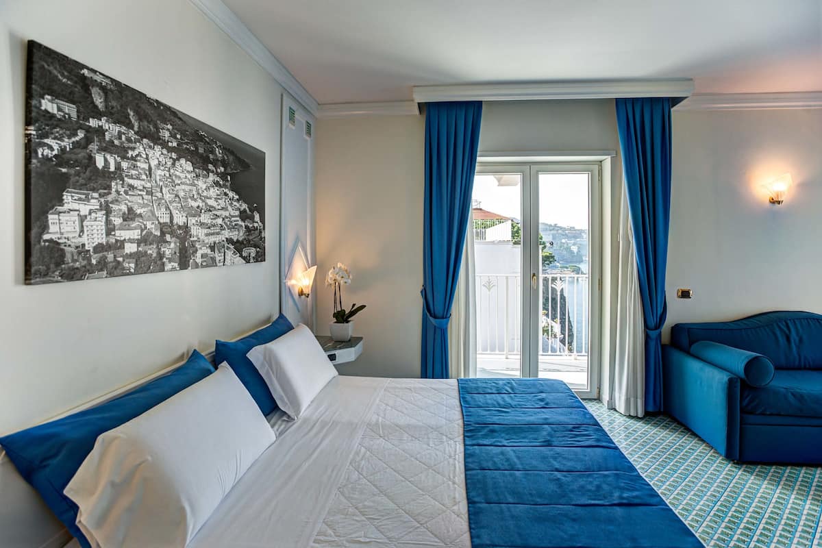 Ein Hotelzimmer mit einem Bett, einem blauen Sofa, einem Balkon mit Blick und einem an der Wand hängenden Bild.