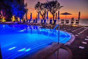 Zona junto a la piscina con sillones y sombrillas, piscina con iluminación azul y vistas al atardecer.