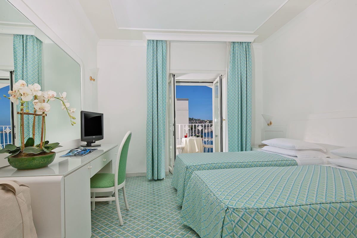 Ein Hotelzimmer mit Schreibtisch, Computer, grünem Sessel, zwei Betten mit passender Bettwäsche, einer Topfpflanze und einem Balkon mit Blick.