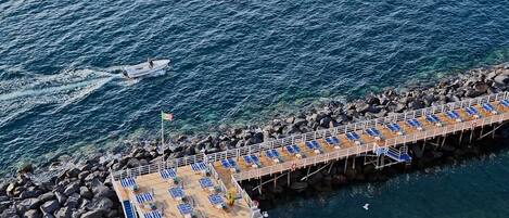 An aerial view of a pier with sun loungers and a flag, extending into a rocky sea.