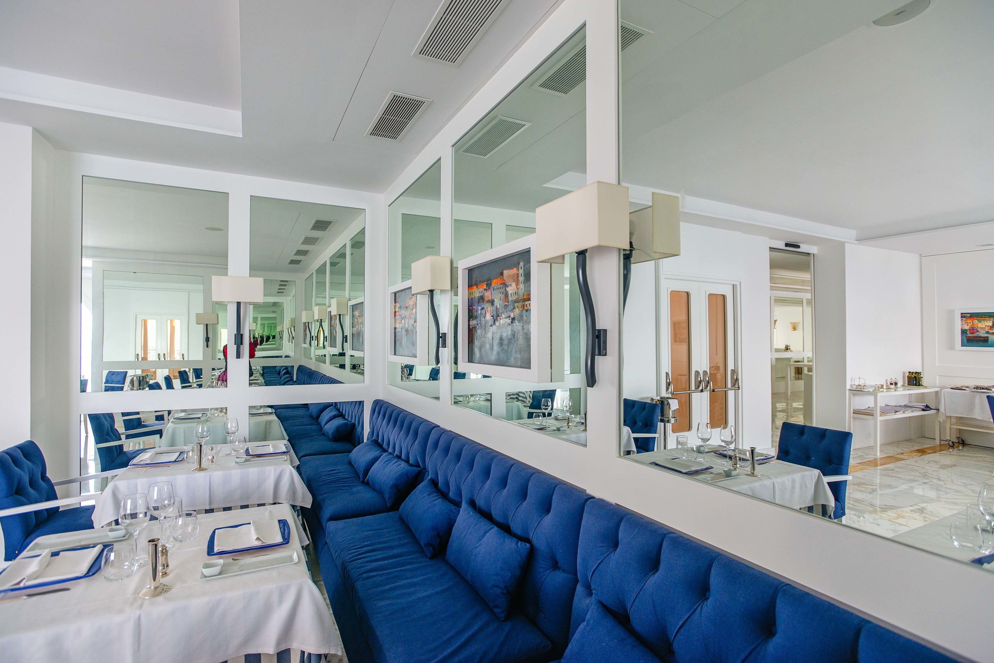 A modern restaurant with blue upholstered seating, white tables, and large mirrors reflecting the interior.