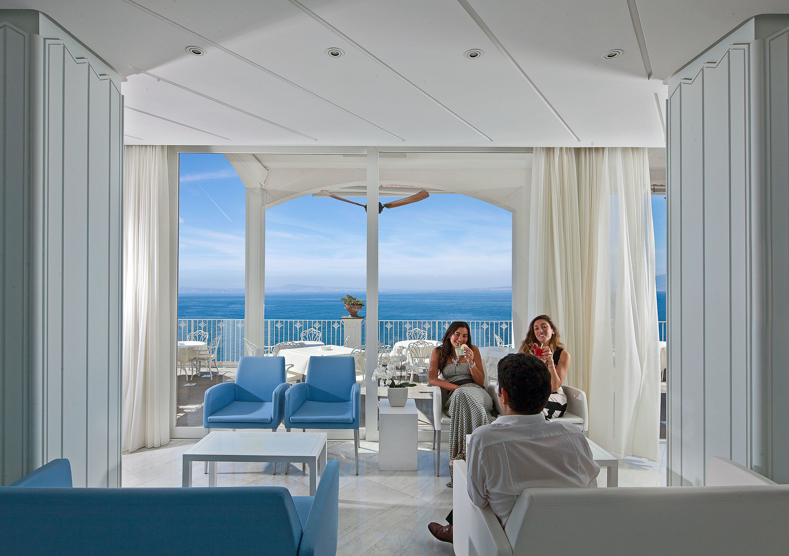 Un balcón espacioso con vistas despejadas al mar, amueblado con sillas azules y mesas blancas, y un techo blanco.